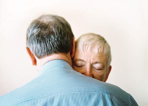 Two elderly individuals embrace closely. The person facing the camera has short gray hair and is smiling gently, while the person with their back to the camera has short gray hair and is wearing a blue and white striped shirt. The background is plain and softly lit.