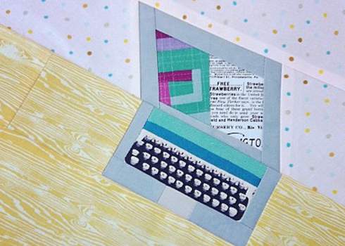 A craft representation of a laptop made from various fabric pieces is shown. The screen is made of newspaper print fabric, while the keyboard has a blue and white pattern. The background includes polka dot and wood grain patterned surfaces.
