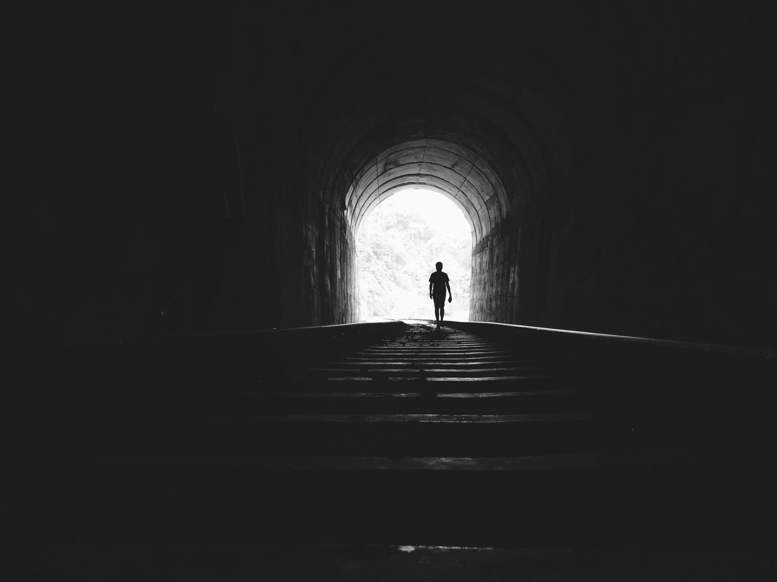 Silhouette of a person walking toward the light at the end of a dark tunnel on railway tracks, symbolizing finding peace.