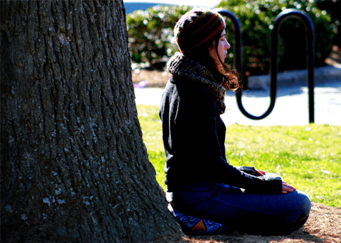 A person wearing a hat and scarf is sitting cross-legged on the ground, leaning against a large tree, outdoors in a sunny area. They appear to be meditating or in a state of relaxation. Blurred greenery and a bike rack are visible in the background.