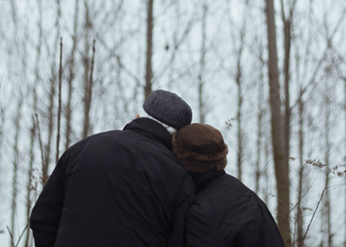 Two individuals, both dressed in dark winter coats and hats, are seen from behind as they lean close to each other, gazing upwards in a forest with leafless trees on a cloudy day. Their close proximity suggests a deeper kind of love and companionship.