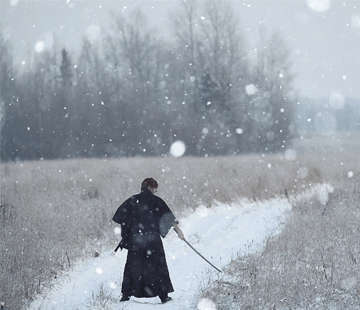 A person dressed in dark, traditional clothing walks down a snow-covered path in a snowy field, holding a long stick or sword. Snowflakes are falling, and bare trees are visible in the background under a gray sky.