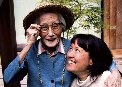 An elderly person wearing a straw hat and glasses smiles while holding the arm of a younger person. The younger person, with dark hair, smiles and looks up affectionately at the elderly person. They are outside near a plant and wooden structure.