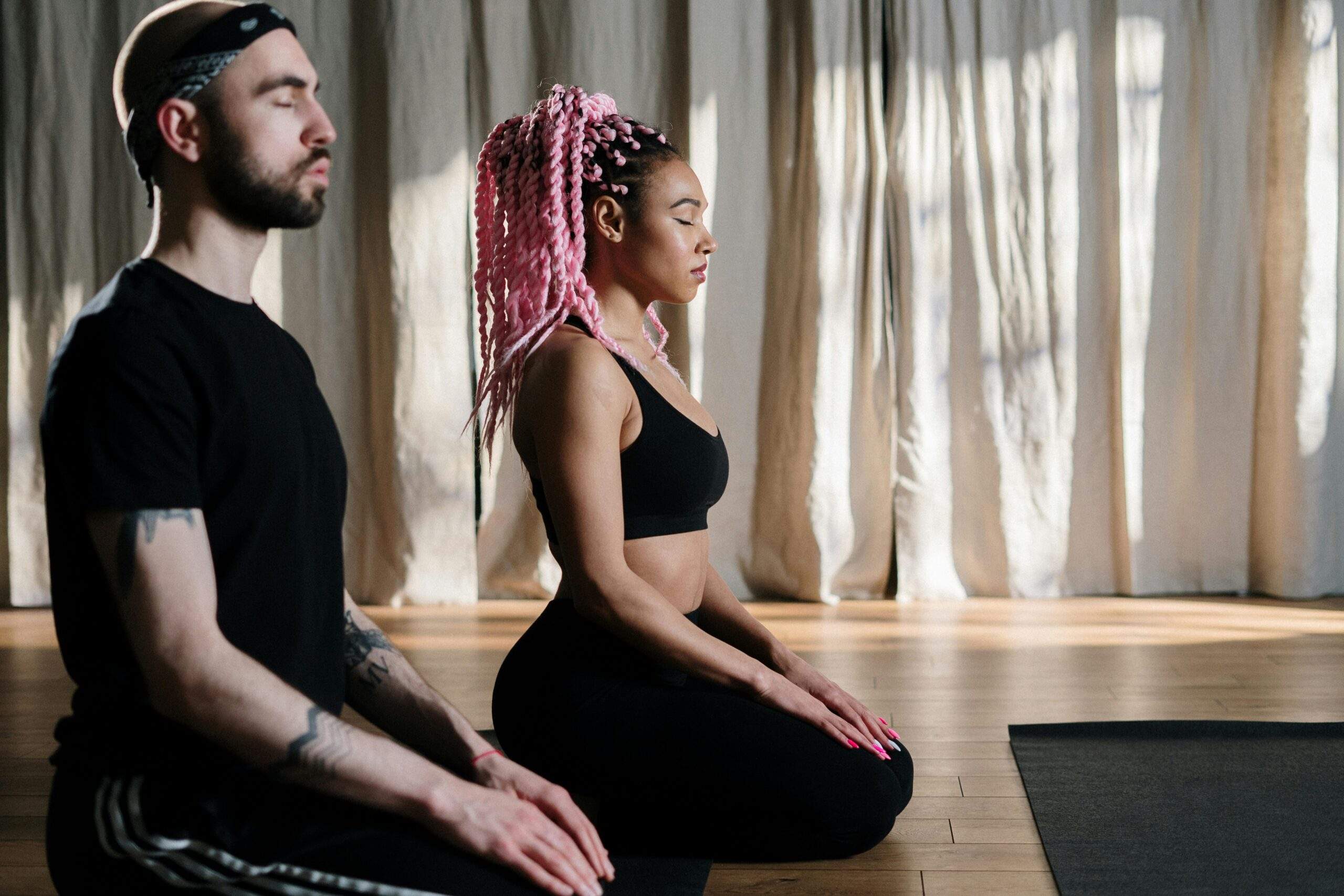 A young man and woman kneel on the floor in a meditation studio with their eyes closed.