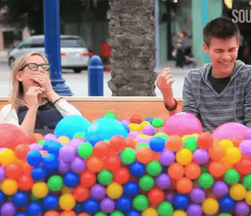 Two people sit on a bench, laughing while surrounded by colorful plastic balls of various sizes. One person wears glasses and holds a hand to their mouth, while the other person smiles with their eyes closed. A palm tree and parked cars are in the background.