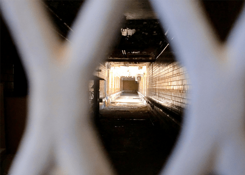 View through a metal fence of a dimly lit, narrow, and long corridor with tiled walls, leading to an area illuminated by bright sunlight at the far end. Debris litters the floor of the corridor, giving it an abandoned feel.