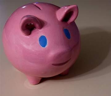 A pink piggy bank with blue eyes and a smile stands on a beige surface. The ceramic piggy bank has a coin slot on its back, embodying the simple and playful style often adored by rich people seeking whimsical decor.