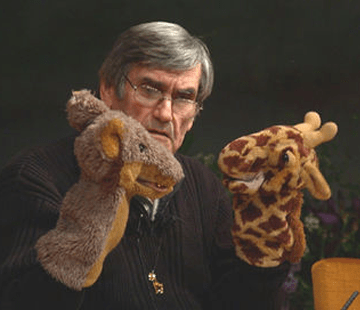 A person with gray hair and glasses is holding up two hand puppets: a gray rodent and a giraffe. The individual looks intently at the puppets, which are positioned as if they are interacting with each other. The background is dark and slightly blurred.