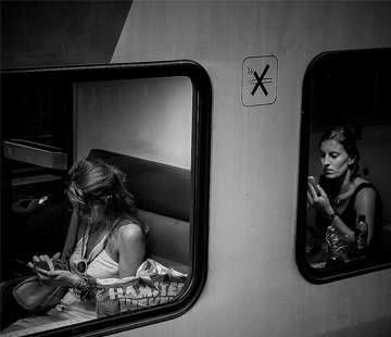 Two women are sitting on a train in separate windows. One is engrossed in her phone, while the other applies makeup using her phone as a mirror. Both windows are separated by a train wall with a no smoking sign. The photo is in black and white.