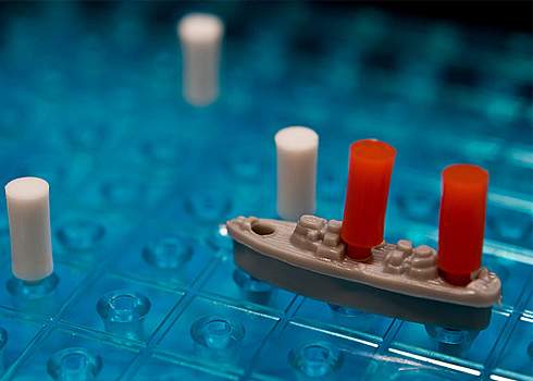 Close-up of a game piece from the board game Battleship. A grey plastic ship with two red pegs is placed on a blue pegboard. Two white pegs are positioned vertically around the ship, indicating potential shots taken in the game.