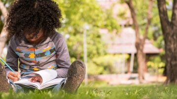 The Surprising Benefits of Teaching a Class Outside A little boy writing on notebook while sitting on green grass in